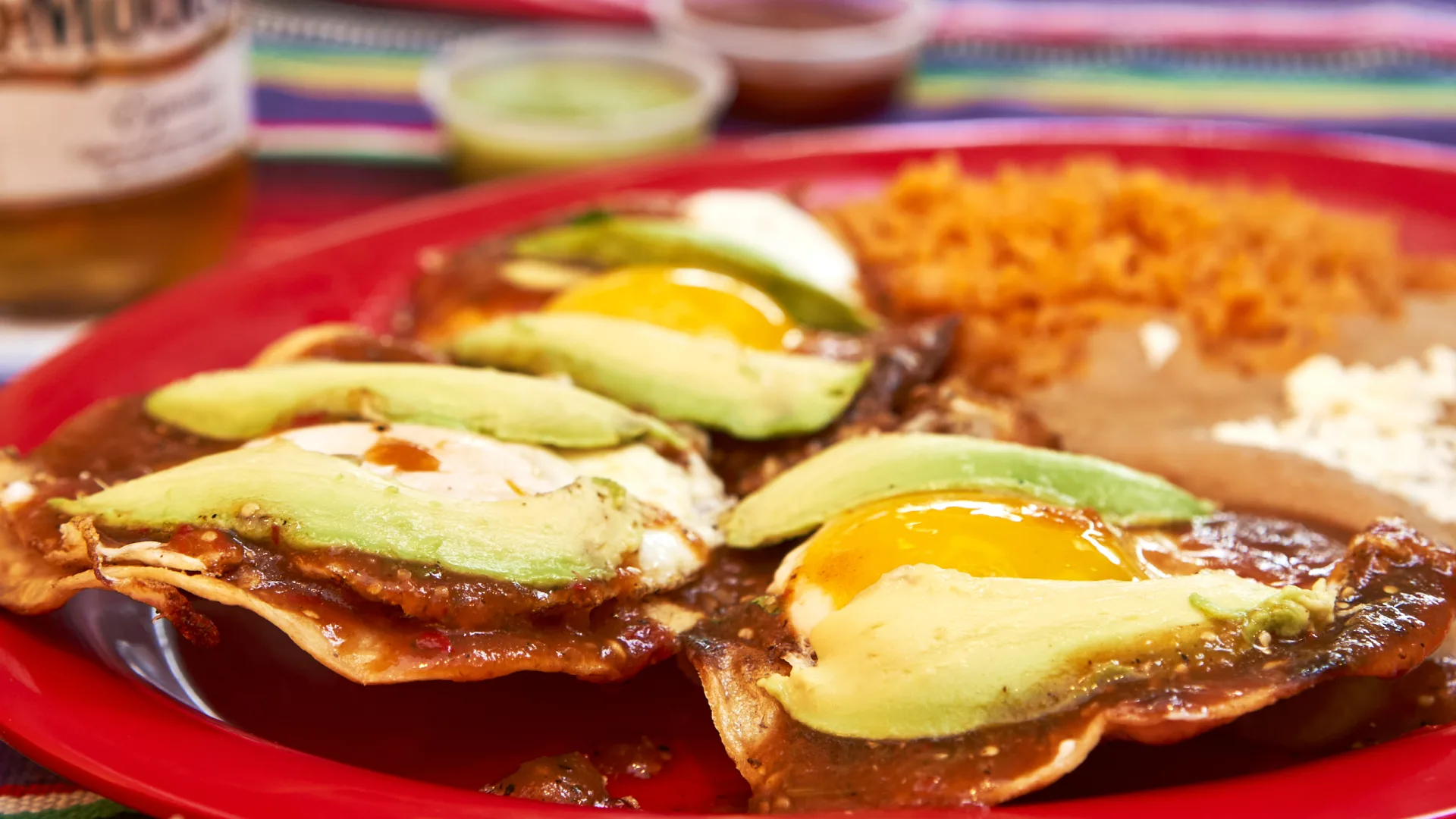 Huevos rancheros with red salsa, avocado, rice and beans on a red plate.